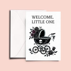 May include: A white greeting card and matching envelope featuring the text WELCOME, LITTLE ONE in black. The card displays a black baby carriage illustration adorned with a skull motif, dark roses, and a small bow, set against a plain white background.