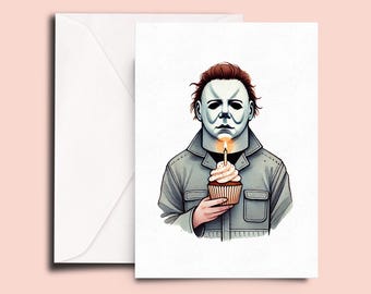 Può includere: Un biglietto d'auguri con l'illustrazione di una figura che indossa una maschera bianca e una giacca grigia con colletto, che tiene in mano un cupcake con una candela accesa. Il biglietto è posizionato davanti a una busta bianca semplice.