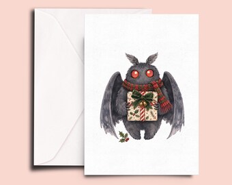 May include: A white greeting card featuring a dark gray, furry moth-like creature with large glowing red eyes. The creature wears a red and green striped scarf and holds a gift box wrapped in holly-patterned paper with a green bow.