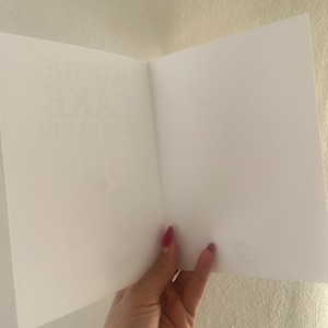 May include: A white greeting card held open to reveal a blank interior. The left side of the card features faint, partially visible text printed in a light gray font that reads "MAY THE CAKE BE WITH YOU" in a vertical orientation.