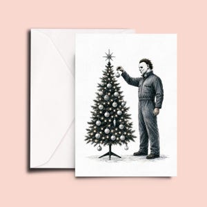Op de afbeelding: Een wenskaart met een zwart-witte inktillustratie van een figuur in een donkere overall en een wit masker die een kerstboom versiert met ronde ornamenten en een ster als piek, vergezeld van een bijpassende witte envelop op een lichte achtergrond.