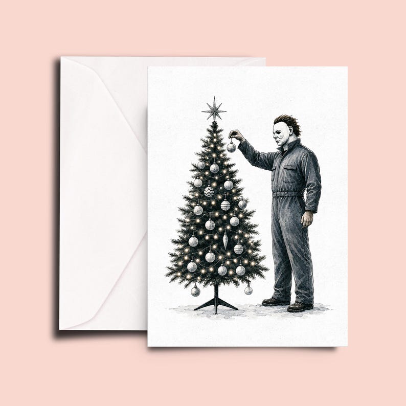 Op de afbeelding: Een wenskaart met een zwart-witte inktillustratie van een figuur in een donkere overall en een wit masker die een kerstboom versiert met ronde ornamenten en een ster als piek, vergezeld van een bijpassende witte envelop op een lichte achtergrond.