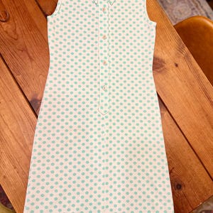 May include: A sleeveless white dress featuring a repeating pattern of small light green polka dots. The dress has a pointed collar, a front button placket with four visible buttons, and a straight hemline. The fabric has a subtle vertical texture.