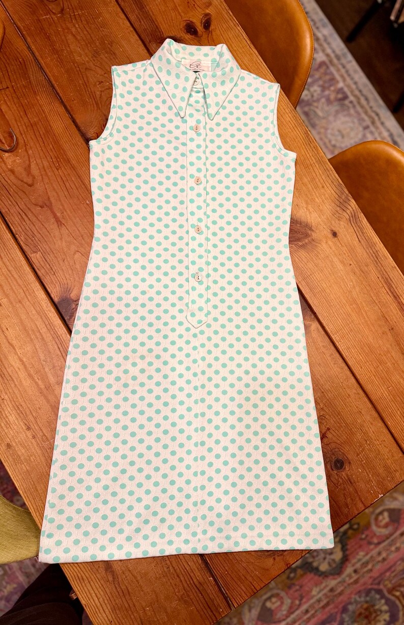 May include: A sleeveless white dress featuring a repeating pattern of small light green polka dots. The dress has a pointed collar, a front button placket with four visible buttons, and a straight hemline. The fabric has a subtle vertical texture.
