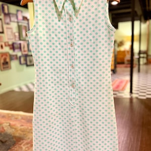 May include: A sleeveless white dress featuring a repeating pattern of light green polka dots. The dress has a pointed collar, a front button-down placket with five visible buttons, and a straight-cut silhouette that falls to the mid-thigh.