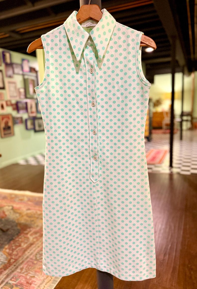 May include: A sleeveless white dress featuring a repeating pattern of light green polka dots. The dress has a pointed collar, a front button-down placket with five visible buttons, and a straight-cut silhouette that falls to the mid-thigh.