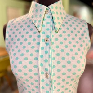 May include: A sleeveless collared shirt featuring a white base with a repeating pattern of light mint green polka dots. The garment includes a front button placket with four visible round buttons and a structured collar, crafted from a textured fabric.