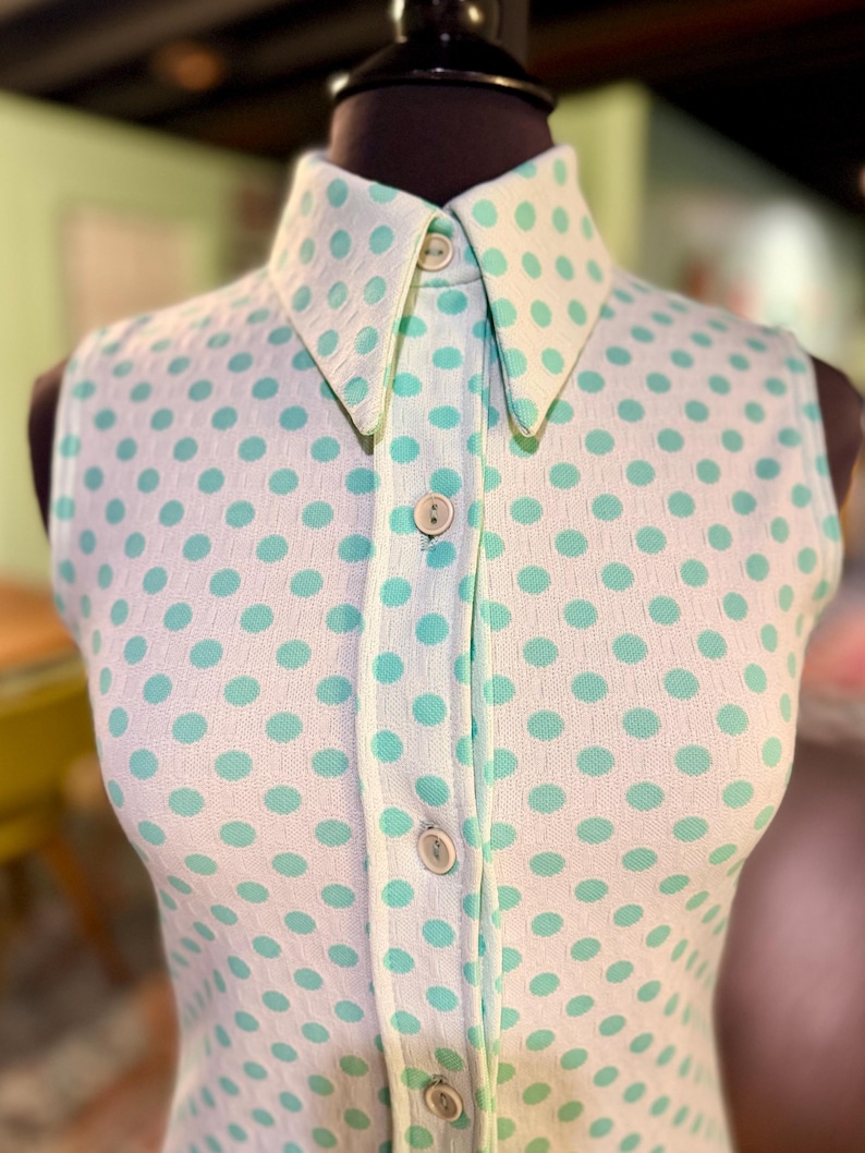 May include: A sleeveless collared shirt featuring a white base with a repeating pattern of light mint green polka dots. The garment includes a front button placket with four visible round buttons and a structured collar, crafted from a textured fabric.