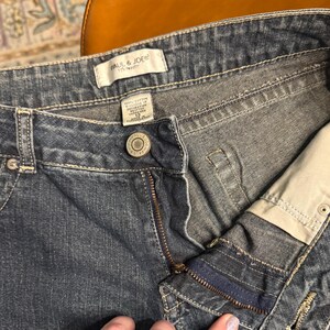 以下が含まれることがあります： 青いデニムジーンズのクローズアップ。ジーンズはボタン留め、ジッパー、ポケットが付いています。「PAUL & JOE」のブランド名がラベルに表示されています。ジーンズのサイズは12です。