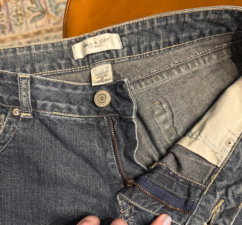 以下が含まれることがあります： 青いデニムジーンズのクローズアップ。ジーンズはボタン留め、ジッパー、ポケットが付いています。「PAUL & JOE」のブランド名がラベルに表示されています。ジーンズのサイズは12です。