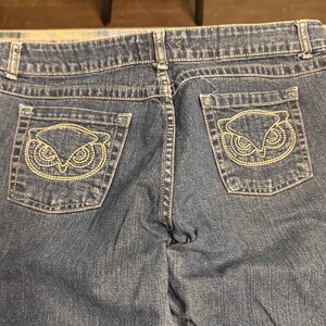 以下が含まれることがあります： 青いデニムジーンズの背面をクローズアップ。ジーンズには2つのバックポケットがあり、それぞれに金色の糸で刺繍されたフクロウのデザインが施されています。ステッチとディテールが見え、衣服の職人技を強調しています。