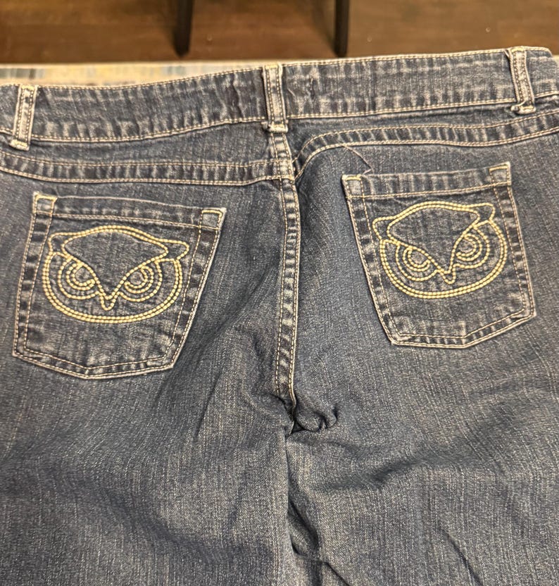 以下が含まれることがあります： 青いデニムジーンズの背面をクローズアップ。ジーンズには2つのバックポケットがあり、それぞれに金色の糸で刺繍されたフクロウのデザインが施されています。ステッチとディテールが見え、衣服の職人技を強調しています。