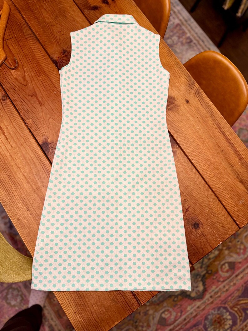 May include: A sleeveless dress featuring a white base with a repeating pattern of small, light green polka dots. The garment has a structured collar at the neckline and a straight, knee-length silhouette, showcasing a clean and simple design.