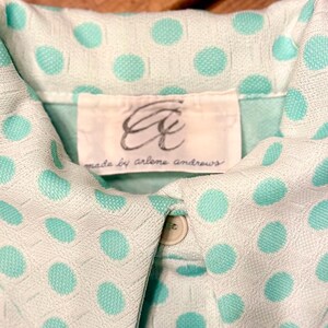 May include: A light mint green collared shirt featuring a textured white polka dot pattern. The garment includes a white interior label with the cursive text "Aa" and "made by arlene andrews" printed in black, along with a single visible beige button.