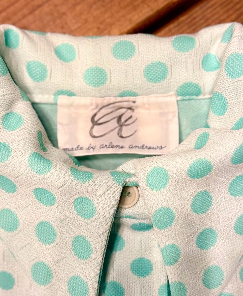 May include: A light mint green collared shirt featuring a textured white polka dot pattern. The garment includes a white interior label with the cursive text "Aa" and "made by arlene andrews" printed in black, along with a single visible beige button.