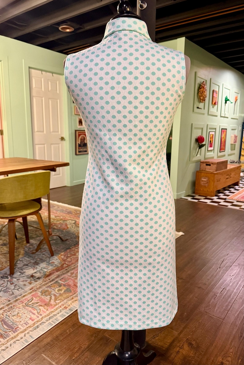 May include: A sleeveless knee-length dress featuring a white base with a repeating pattern of light teal polka dots. The garment includes a structured collar at the neckline and a straight-cut silhouette, displayed on a mannequin form.
