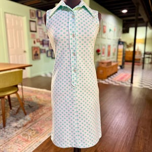 May include: A sleeveless knee-length dress featuring a white base with a repeating pattern of small light green polka dots. The dress has a pointed collar, a partial front button placket with four visible buttons, and a straight-cut hemline.