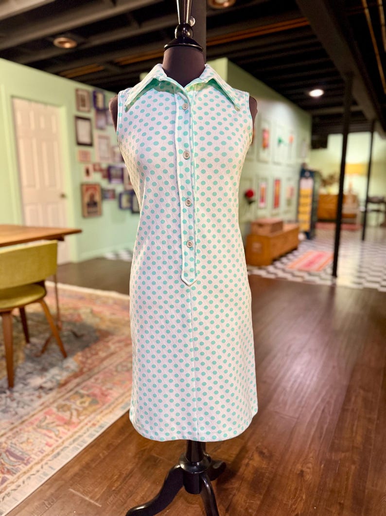 May include: A sleeveless knee-length dress featuring a white base with a repeating pattern of small light green polka dots. The dress has a pointed collar, a partial front button placket with four visible buttons, and a straight-cut hemline.