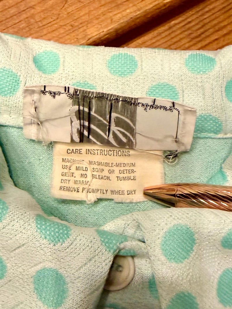 May include: A light mint green garment with a textured polka dot pattern. A white care label is attached, featuring the text: CARE INSTRUCTIONS, MACHINE WASHABLE-MEDIUM, USE MILD SOAP OR DETERGENT, NO BLEACH, TUMBLE DRY WARM, REMOVE PROMPTLY WHEN DRY.
