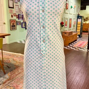 May include: A sleeveless white dress featuring a repeating pattern of light green polka dots. The dress has a pointed collar, a central button-down placket with five visible buttons, and a straight-cut silhouette that falls to the knee.