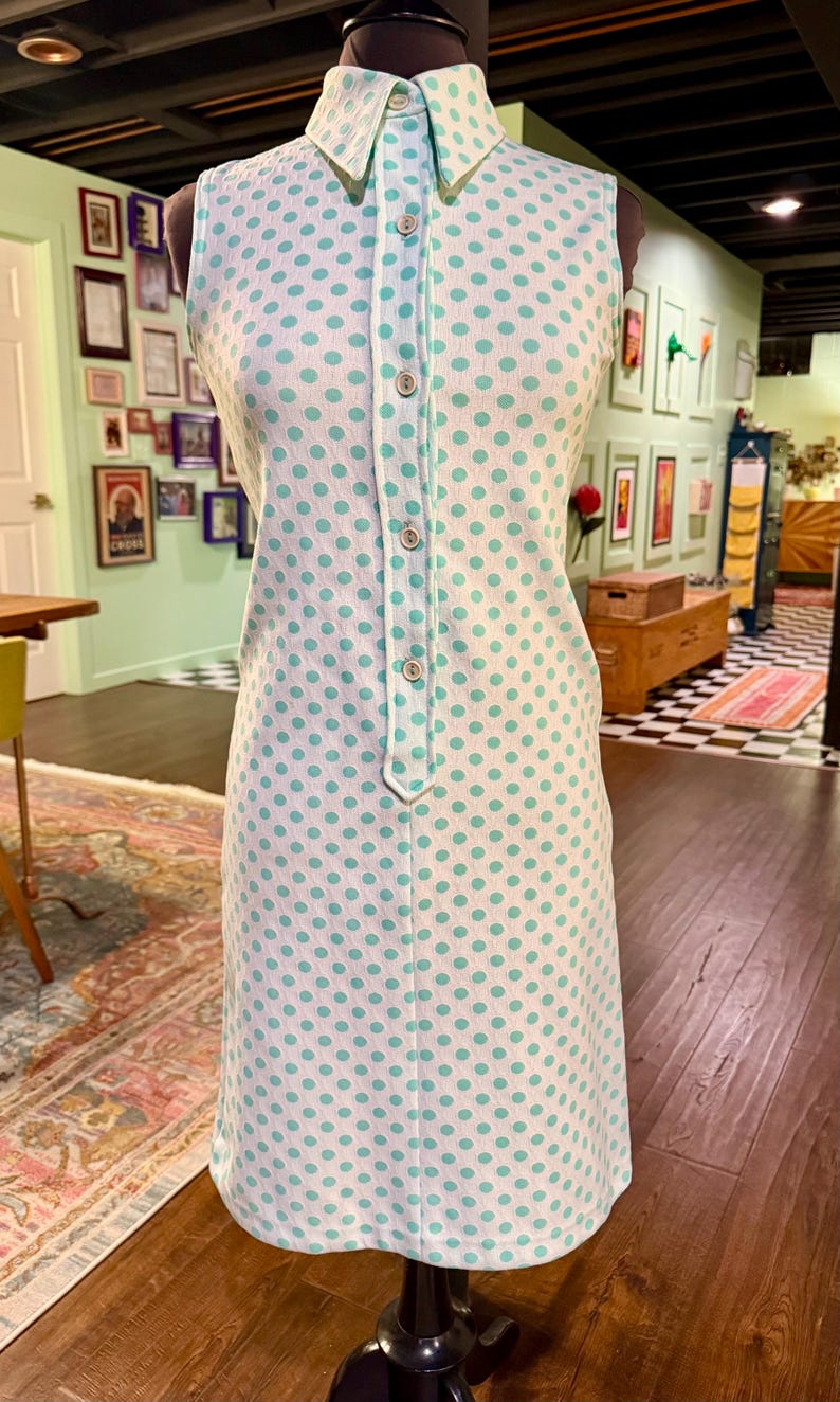May include: A sleeveless white dress featuring a repeating pattern of light green polka dots. The dress has a pointed collar, a central button-down placket with five visible buttons, and a straight-cut silhouette that falls to the knee.
