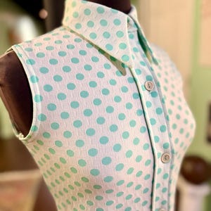 May include: A sleeveless collared shirt featuring a white base with a repeating pattern of light turquoise polka dots. The garment includes a front button placket with four visible round buttons and a textured, grid-like fabric construction.