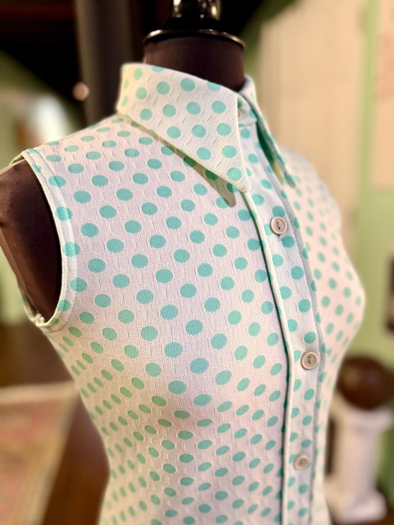 May include: A sleeveless collared shirt featuring a white base with a repeating pattern of light turquoise polka dots. The garment includes a front button placket with four visible round buttons and a textured, grid-like fabric construction.