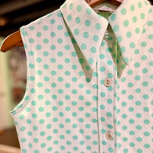 May include: A sleeveless collared shirt featuring a white base with a repeating mint green polka dot pattern. The garment includes a pointed collar, a front button placket with three visible light-colored buttons, and a textured fabric finish.