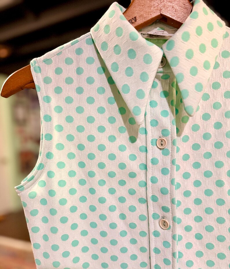 May include: A sleeveless collared shirt featuring a white base with a repeating mint green polka dot pattern. The garment includes a pointed collar, a front button placket with three visible light-colored buttons, and a textured fabric finish.