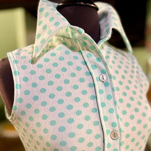 May include: A sleeveless collared shirt featuring a white base with a repeating pattern of light green polka dots. The garment has a button-down front placket with two visible round buttons and a structured collar, crafted from a textured fabric.