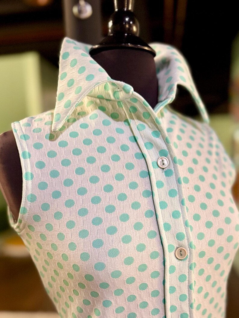 May include: A sleeveless collared shirt featuring a white base with a repeating pattern of light green polka dots. The garment has a button-down front placket with two visible round buttons and a structured collar, crafted from a textured fabric.