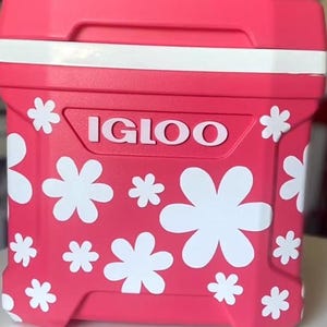 May include: A bright pink Igloo cooler featuring a white horizontal band near the top and a pattern of white flower decals across the front. The brand name IGLOO is embossed in white capital letters in the center of the front panel.