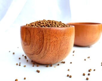 May include: Two wooden bowls filled with coriander seeds. The bowls are on a white surface with scattered coriander seeds.