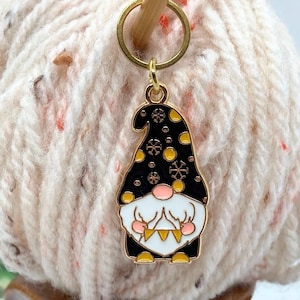 Könnte beinhalten: Ein goldfarbener Maschenmarkierer in Zwergform mit einem schwarzen Hut, der mit gelben Kreisen und Schneeflocken verziert ist. Der Zwerg hat einen weißen Bart, eine rosa Nase und einen goldfarbenen Körper. Der Maschenmarkierer ist an einem goldenen Ring befestigt und auf einem Knäuel Wolle platziert.