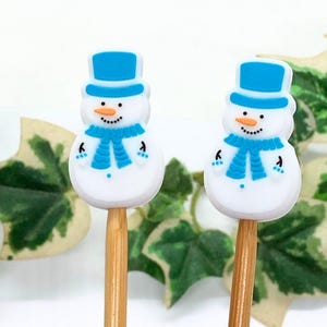Könnte beinhalten: Zwei dekorative Zahnstocher in Form von Schneemännern. Jeder Schneemann hat einen weißen Körper, einen blauen Hut und Schal sowie eine Karottennase. Die Zahnstocher sind auf Holzstäbchen, ideal für Winterdekorationen.