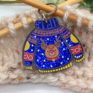 Könnte beinhalten: Ein blauer, pulloverförmiger Schlüsselanhänger mit Rentier-Design. Der Schlüsselanhänger hat einen glitzernden blauen Hintergrund mit einem Rentiergesicht in der Mitte. Der Pullover hat gelbe Akzente und rote Herzformen. Stricknadeln und Garn im Hintergrund.
