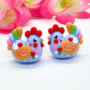Könnte beinhalten: Ein Paar hellblaue, huhnförmige Ohrringe. Jeder Ohrring hat einen roten Kamm, einen orangefarbenen Schnabel und bunte Schwanzfedern. Die Hühner sind mit Herzen und Punkten verziert. Rosa Blumen sind im Hintergrund.