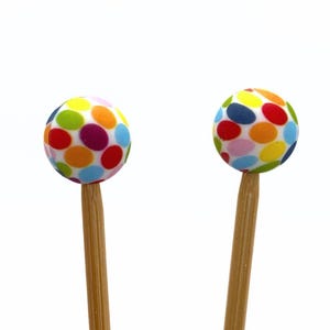Könnte beinhalten: Zwei dekorative Cocktailspieße. Jeder Spieß hat eine Kugel mit einem farbenfrohen Tupfenmuster in Rot, Orange, Gelb, Grün, Blau und Lila auf einem Holzspieß. Der Hintergrund ist weiß.