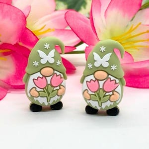 Könnte beinhalten: Zwei skurrile Zwergenfiguren. Jeder Zwerg trägt einen grünen Hut mit weißen Schmetterlings- und Sternakzenten. Sie halten rosa Tulpensträuße. Die Zwerge haben weiße Körper, hellbraune Nasen und schwarze Schuhe. Rosa und gelbe Blumen sind im Hintergrund.