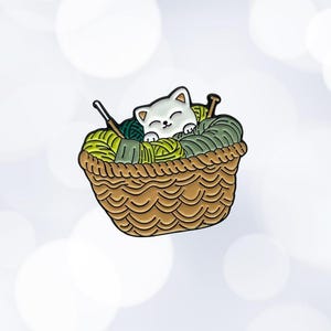 Könnte beinhalten: Eine weiße Katze schläft in einem braunen Weidenkorb, der mit grünem und weißem Garn gefüllt ist. Die Katze ist von Garn und einer Stricknadel umgeben.