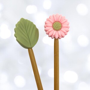 Könnte beinhalten: Zwei Stricknadeln aus Holz mit dekorativen Spitzen. Eine Nadel hat eine rosa Blütenspitze und die andere eine grüne Blattspitze.