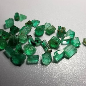 Può includere: Una collezione di cristalli di smeraldo grezzi, di varie dimensioni e forme. Le pietre preziose mostrano una vibrante tonalità verde, con alcune che presentano inclusioni e texture naturali. I cristalli sono sparsi su una superficie chiara, mettendo in risalto la loro bellezza naturale.