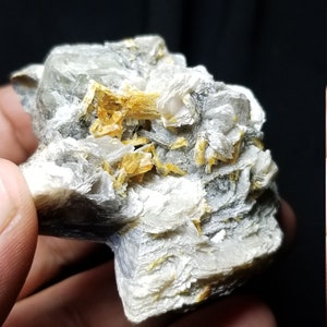 May include: A close-up of a mineral specimen held in a hand. The rock has a layered, crystalline structure with shades of gray, white, and yellow. The mineral appears to be a geological sample.