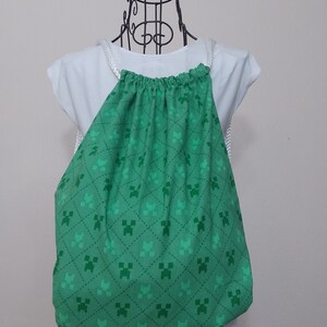 May include: Green drawstring backpack with a repeating pattern of pixelated green characters. The bag is made of fabric and has white rope straps. The bag is displayed on a white mannequin.