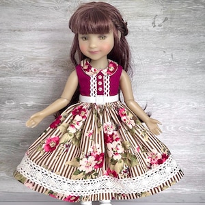 May include: A doll in a burgundy and floral dress with a white lace trim. The dress features a white collar and belt. The doll has brown hair and wears white shoes. The dress has a striped pattern.