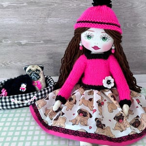 May include: A handmade doll with long brown hair, green eyes, and a pink knit hat and sweater. The doll wears a skirt patterned with pug dogs. A small pug dog toy rests in a black and white checked bed, with the text "Pug Love".