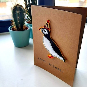 Verjaardagskaart met papegaaiduiker - uit de vrije hand geborduurde papegaaiduiker met een feestmuts, gemonteerd op een bruine A6-kaart van kraftpapier.