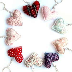 Op de afbeelding: Een verzameling van tien hartvormige sleutelhangers met verschillende stoffen patronen. De sleutelhangers zijn in een cirkel op een witte achtergrond gerangschikt. De patronen omvatten bloemen, stippen, ruitjes en effen kleuren.