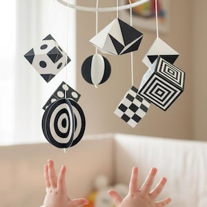 Pode incluir: Móvel de bebê em preto e branco com formas geométricas suspensas de uma estrutura circular branca. As formas incluem cubos, esferas e desenhos planos com padrões. O móvel está pendurado acima de um berço.
