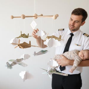 Puede incluir: Móvil para bebé de madera con aviones de papel y nubes blancas suspendidos de un marco de madera. Los aviones son de varios colores y diseños. Diseñado para una guardería.